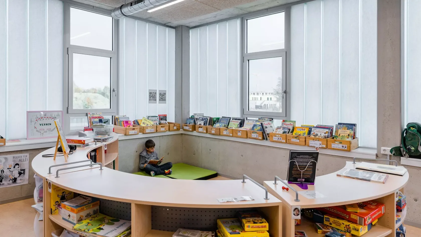 Ein Blick in ein Lesezimmer, im Vordergrund ein halbrundes Regal, gefüllt mit Büchern und Spielzeug, im Hintergrund an der Wand sitzend ein kleiner Junge mit Buch in der Hand.