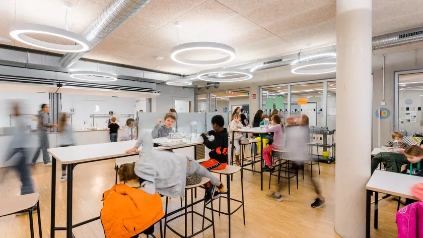 Ein Blick in einen Klassenraum, Kinder und Lernbegleiter:innen stehend und sitzend an Hochtischen.